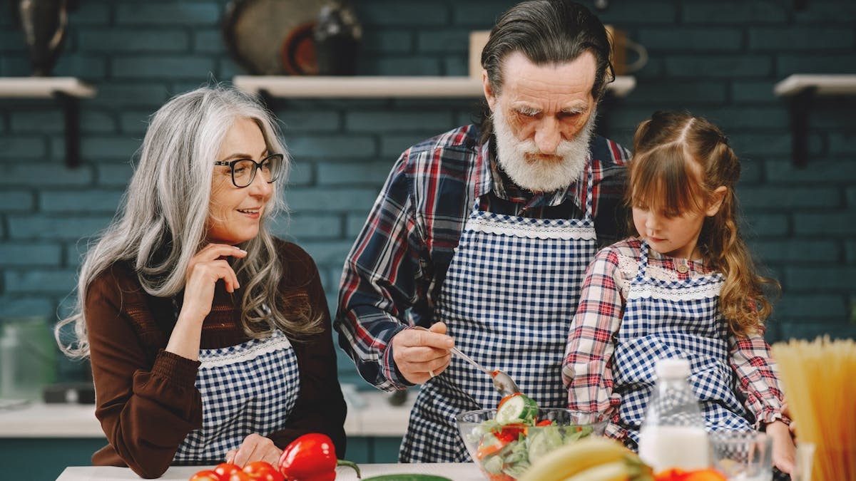 Grandparents and granddaughter preparing salad in home kitchen, creating memories together.