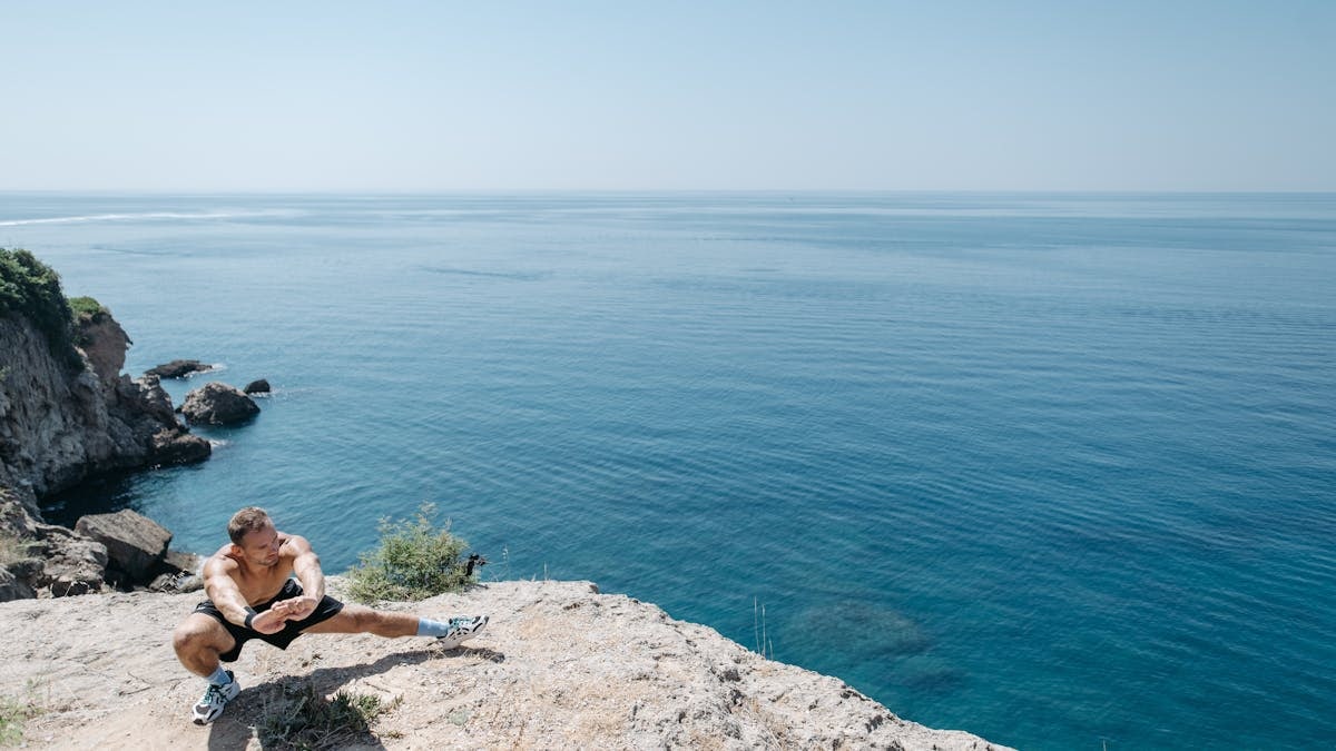 Shirtless man stretching on a cliff's edge by the ocean showcasing a healthy lifestyle and fitness.